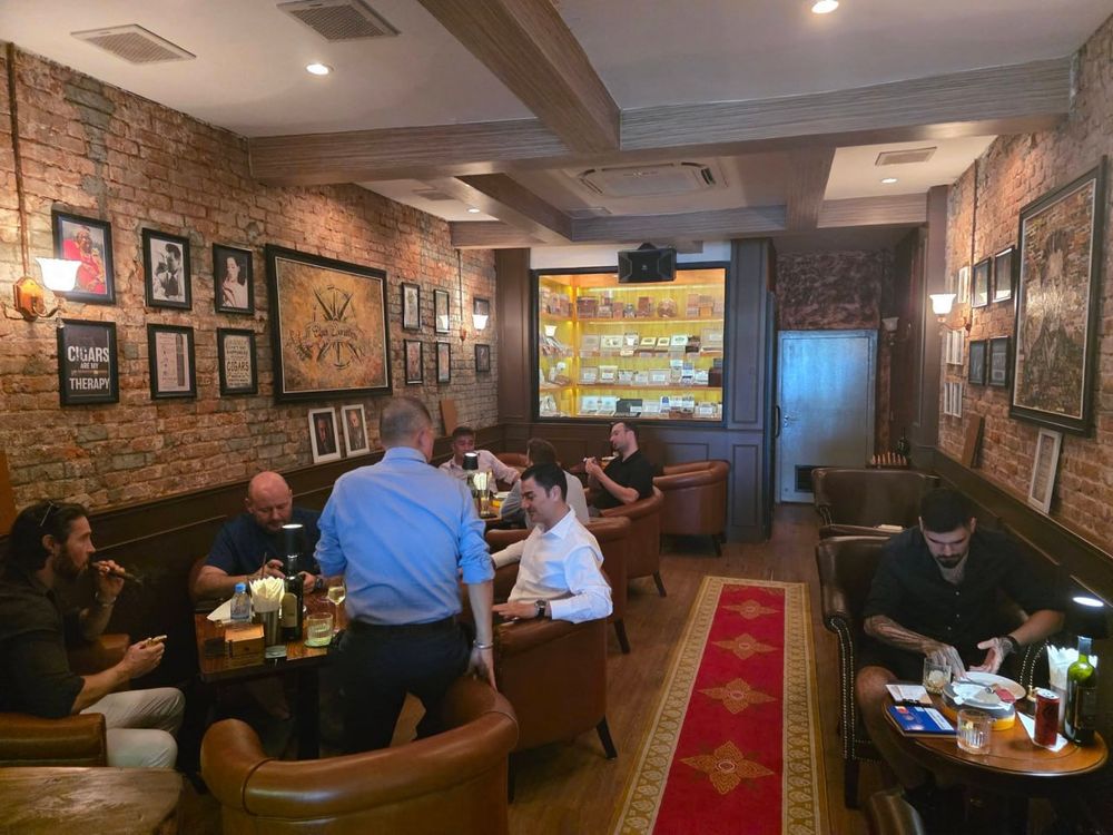 The Cigar Connection lounge in the evening, guests smoking cigars in leather armchairs with the humidor lit at the back of the room
