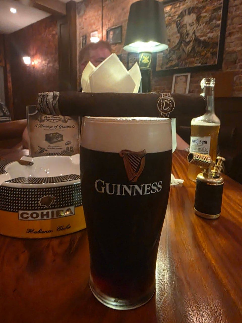A pint of Guinness on a wooden table beside a large cigar resting on a Cohiba ashtray