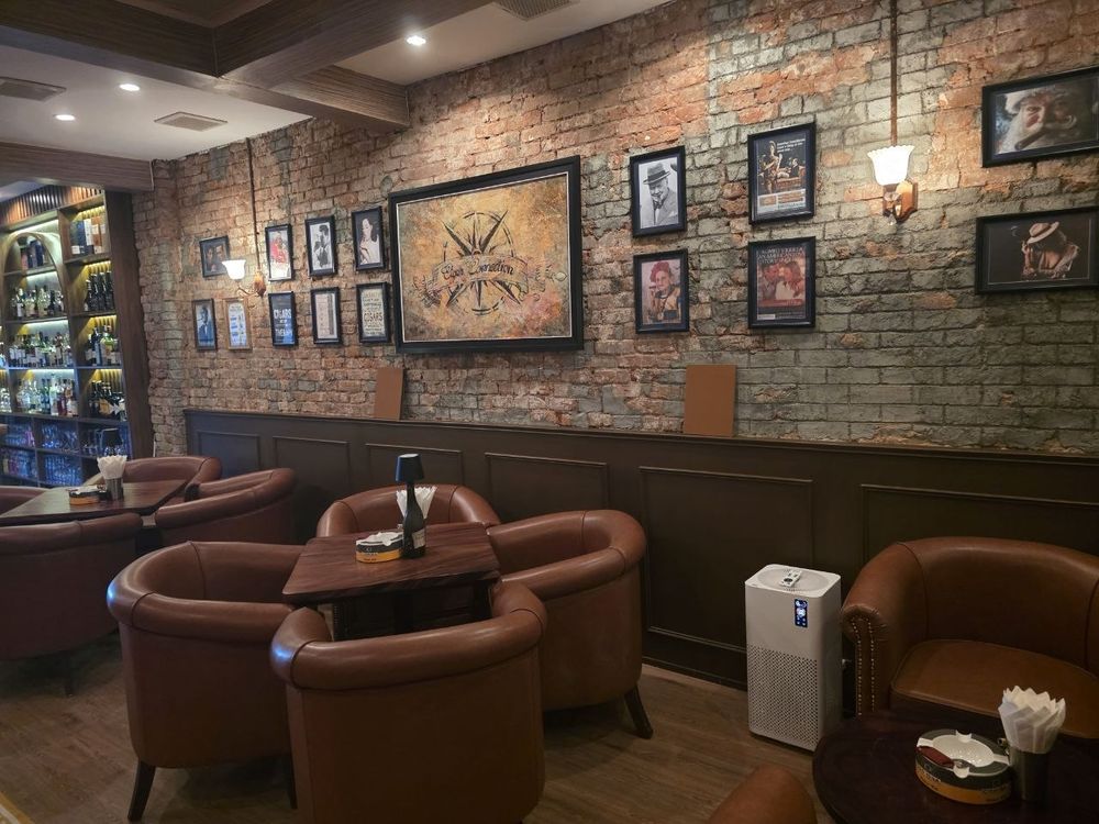 The brick wall of the lounge hung with framed vintage cigar artwork and portraits, with leather armchairs below and a lit bar to one side